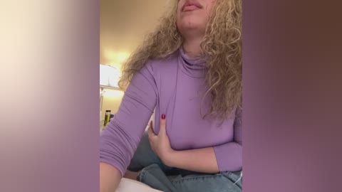 Media: Video of a smiling, light-skinned woman with curly blonde hair, wearing a lavender long-sleeve top and blue jeans, seated on a couch with a warm, lit room in the background.