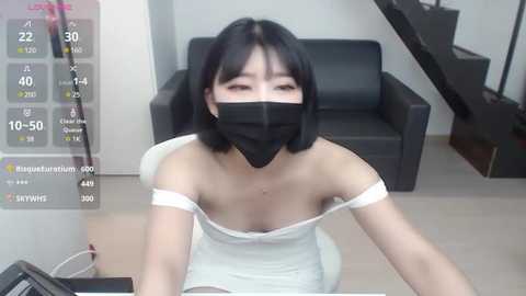 Media: Video of an Asian woman with shoulder-length black hair, wearing a black face mask, white off-shoulder dress, and sitting on a white chair in a minimalist room with a black couch and a digital display.