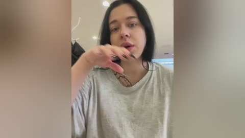 Media: Video of a young woman with straight black hair, wearing a gray T-shirt, in a room with a white ceiling and a visible window. She is holding a black object near her mouth.