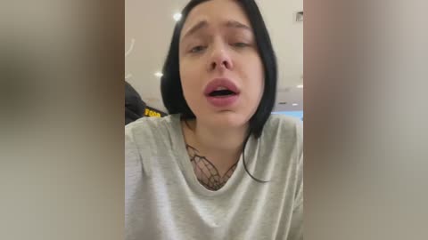 Media: A video of a young woman with fair skin and straight black hair, wearing a grey t-shirt, looking surprised with her mouth open, blurred background.