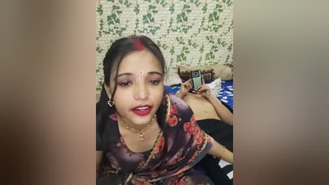 Media: Video of a young woman with dark skin and long black hair, adorned with red mehndi and bindi, wearing a floral saree, lying on a bed with blue and white bedding, in a room with green ivy wallpaper.