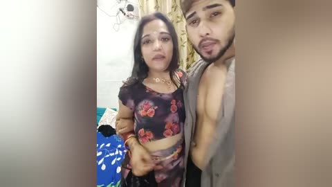 Media: Video of a South Asian woman with long black hair and a floral-patterned top, and a young man with a beard and a grey shirt, standing in a dimly lit room with a bed and curtains in the background.