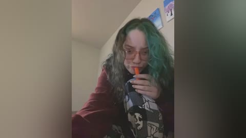 Media: A video of a young woman with long, wavy hair dyed teal and brown, wearing glasses, a patterned scarf, and a maroon jacket, taking a selfie with an orange straw in a white room.