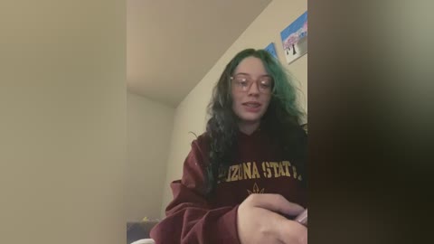 Media: Video of a young woman with long, curly green hair and glasses, wearing a maroon hoodie with \"IZONA STATE\" in gold letters, seated indoors with a beige wall and framed picture in the background.