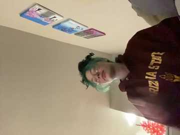 Media: A video of a smiling woman with green hair and glasses, wearing a maroon University of Wisconsin sweatshirt, standing in a beige room with framed artwork on the wall.