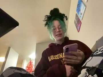 Media: Video of a young person with dyed teal hair in a messy bun, wearing a dark maroon hoodie with \"Iowa\" printed in yellow, sitting on a bed, holding a pink smartphone.