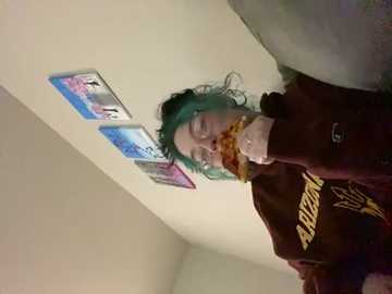 Media: A video shows a person with green hair eating a slice of pizza, wearing a maroon hoodie with \"UVA\" in yellow on the chest. The background features a white wall with two framed pictures.