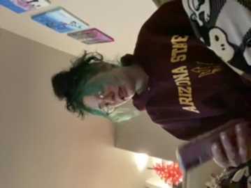 Media: Video of a young person with green hair, wearing a dark maroon Arizona State University t-shirt, standing in a dimly lit room with colorful posters and a red plant.