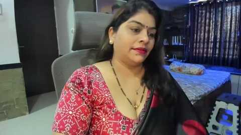 Media: A video of a South Asian woman with medium complexion, long dark hair, and traditional red saree with black border, sitting in a modern bedroom with a gray sofa, wooden blinds, and a bed.