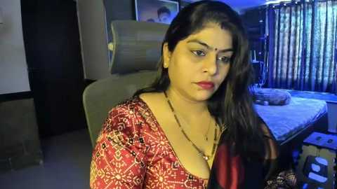 Media: Video of a woman with medium skin tone, long black hair, wearing a red patterned saree and gold jewelry, sitting in a dimly lit room with a bed and window with vertical blinds.