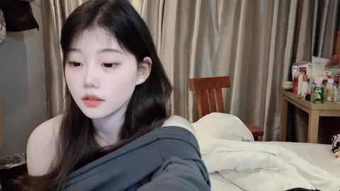 Media: A video of an East Asian woman with long black hair, wearing a gray jacket, sitting on a bed with white sheets, in a dimly lit room with beige curtains and wooden furniture.