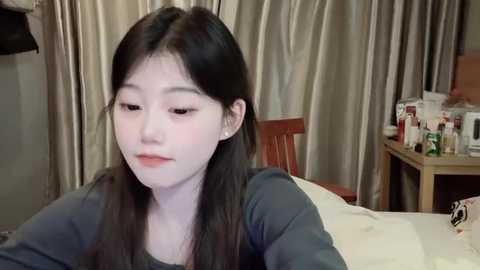 Media: A video of a young East Asian woman with fair skin and long black hair, wearing a dark jacket, sitting on a bed in a bedroom with beige curtains and a wooden table cluttered with items.