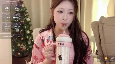 Media: Video of a young East Asian woman in a pink kimono, drinking from a white and pink straw cup, in a cozy indoor setting with a decorated Christmas tree in the background.