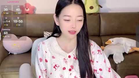 Media: A video of an East Asian woman with long black hair, wearing a white pajama with red animal prints, sitting on a brown couch. Background includes stuffed animals and a screen displaying a health app.