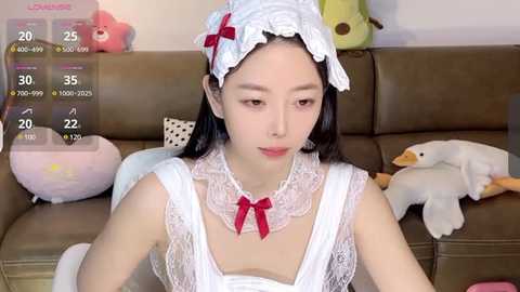 Media: Video of an East Asian woman in a white maid costume with a lace headpiece, sitting on a brown couch surrounded by plush toys, including a duck.