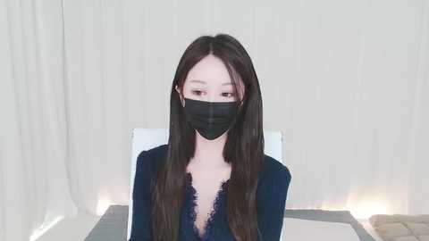 Media: A video of a young Asian woman with long, straight black hair, wearing a black mask, a black lace top, and sitting on a white chair against a white curtain backdrop.