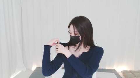 Media: Video of an East Asian woman with long dark hair, wearing a black mask and navy blue long-sleeved top, adjusting her hair in a minimalist, softly lit room with white curtains and a reflective surface.