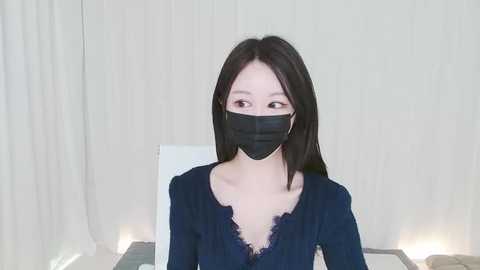 Media: Video of an East Asian woman with long black hair, wearing a black face mask and navy blue lace-trimmed blouse. She stands in a softly lit room with white curtains and a bed in the background.