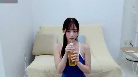 Media: A video of an Asian woman with long black hair, wearing a blue tank top, drinking beer from a can in a minimalist, white-walled room with a beige couch and a small table.