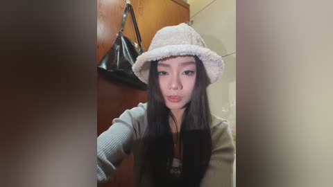 Media: A video of an Asian woman with long black hair, wearing a beige floppy hat, black top, and grey cardigan, standing in a bathroom with a wooden door and a black handbag hanging on it.