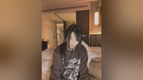 Media: A blurry video of a young Asian woman with long black hair, wearing a dark hoodie, seated in a modern, dimly-lit bedroom with white bedding and wooden furniture.