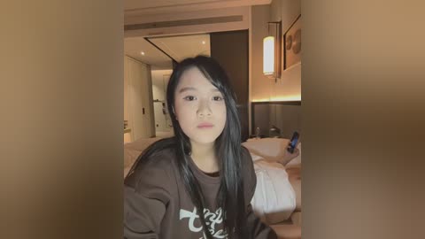 Media: Video of an Asian woman with long black hair, wearing a dark brown T-shirt, standing in a modern, dimly-lit bedroom with white bedding, wooden headboard, and warm lighting.