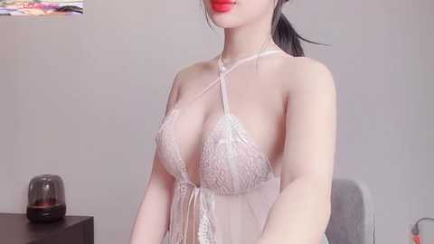 Media: Video of an East Asian woman with fair skin, wearing a sheer, lace, white lingerie top, revealing her cleavage, with red lipstick. She stands against a plain white wall in a minimalist room with a grey chair and a black speaker in the background.