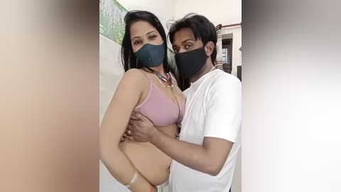 Media: Video of a South Asian woman in a pink bra and black mask, being held by a man in a white shirt and black mask. They are indoors with light-colored walls and greenery visible in the background.