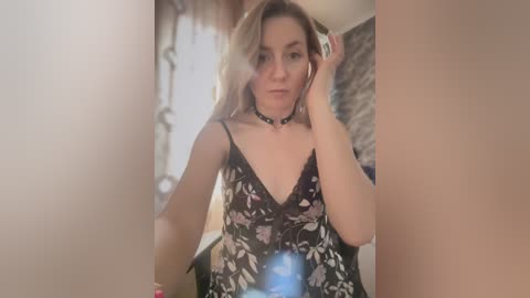 Media: Video of a fair-skinned woman with blonde hair, wearing a black floral camisole, black choker, and headphones, adjusting her hair in a bathroom with beige walls and a mirror.