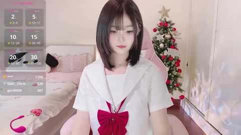 Media: Video of a young Asian woman with straight black hair, wearing a white sailor school uniform with a red bow, sitting on a bed. Background features a Christmas tree and a weather app overlay.