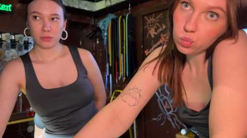 Media: Video of two women in a dimly lit room. One has fair skin, dark hair, and a grey tank top; the other, with fair skin, red hair, and a grey tank top, puckers her lips.