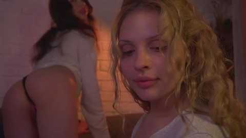 Media: Video of a blonde woman with curly hair, wearing a white sweater, standing in a dimly lit room with another woman in the background, leaning against a brick wall.