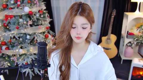 Media: Video of a young woman with long, wavy auburn hair, wearing a white hoodie, singing into a microphone in a cozy, festive room with a decorated Christmas tree and a guitar.