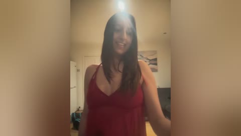 Media: A video of a smiling woman with long brown hair, wearing a red tank top, standing in a dimly lit room with beige walls and a closed door.