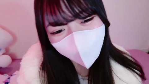 Media: A video of a young Asian woman with long, straight black hair, wearing a white face mask and a white fur-trimmed jacket. Background features soft pastel colors and a plush toy.