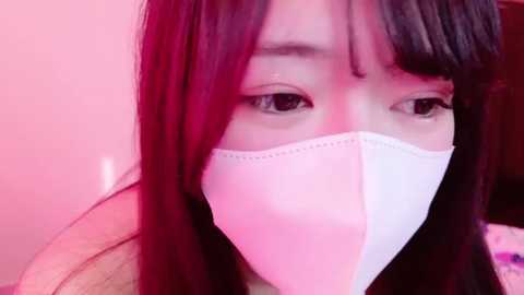 Media: Video of an Asian woman with long black hair, wearing a white surgical mask, in a pink-lit room. Her eyes are partially visible, looking forward.