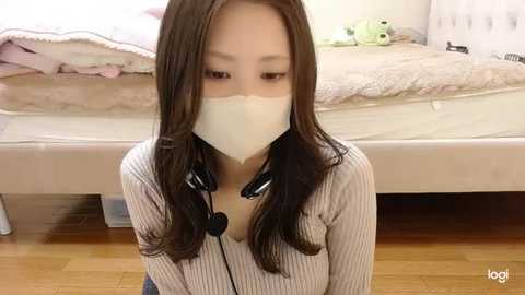 Media: Video of an Asian woman with long brown hair, wearing a beige ribbed sweater, a white face mask, and black headphones, kneeling on a wooden floor in a bedroom with a neatly made bed and stuffed animals.