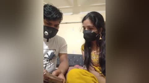 Media: Video: A young man in a white T-shirt with a mask, assisting a woman in a yellow sari with a mask. They're seated indoors.