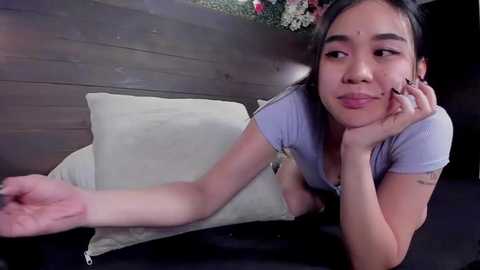 Media: Video of an Asian woman with long black hair, wearing a light blue shirt, lying on a dark couch, smiling while resting her chin on her hand.