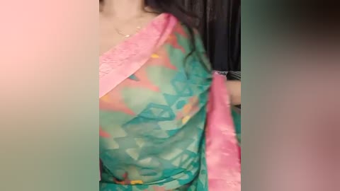 Media: Video of a person wearing a vibrant, semi-transparent sari with a pink border and teal, green, and orange floral pattern. The background is blurred and dark, featuring a hint of a black curtain.