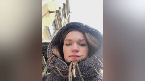 Media: Video of a young Caucasian woman with light skin, wearing a dark, furry hood with long, stringy hair hanging out. The background is blurred, showing a wooden structure and greenery.