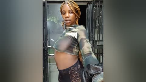 Media: Video of a young Black woman with light skin, long braided hair, and a camo crop top, standing in front of a black iron gate.