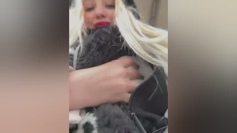 Media: Video of a blonde woman with fair skin and red lipstick, wrapped in a gray, furry blanket, holding a fluffy cat. Background is blurred.