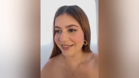 Media: Video of a smiling, fair-skinned woman with long brown hair, wearing minimal makeup and a simple gold necklace, against a blurred background.