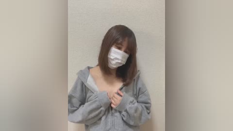 Media: Video of an East Asian woman with shoulder-length brown hair, wearing a gray hoodie, a white face mask, and a gray mask. She stands in a narrow, beige-walled hallway, partially obscured by the hoodie's open front.