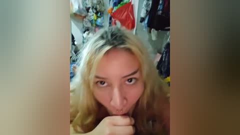 Media: Video of a blonde woman with a nose ring, wearing a top, in a cluttered bedroom with clothes and bags in the background.