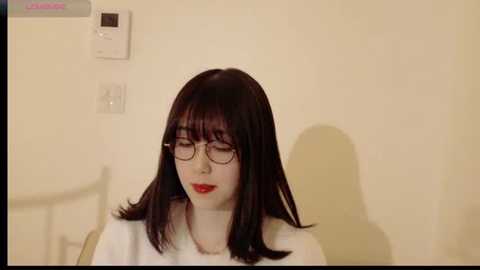 Media: Video of a pale-skinned woman with straight, shoulder-length dark brown hair, wearing red lipstick, black-rimmed glasses, and a white robe, seated indoors against a cream-colored wall with a thermostat and light switch visible.