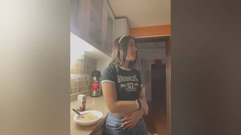 Media: Video of a young woman with long, dark hair in a black \"Brooklyn\" t-shirt, standing in a kitchen with tiled backsplash, wooden floor, and a red coffee maker.