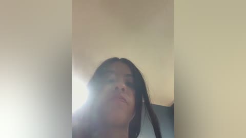 Media: Video of a young woman with straight, dark hair and medium skin tone, captured from a low angle, partially obscured by lens flare. She has a neutral expression and is indoors against a beige wall.