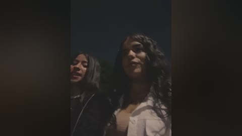 Media: A dimly lit video featuring two young women with wavy hair, one with a black jacket and the other in a white shirt. Background is dark and blurry.